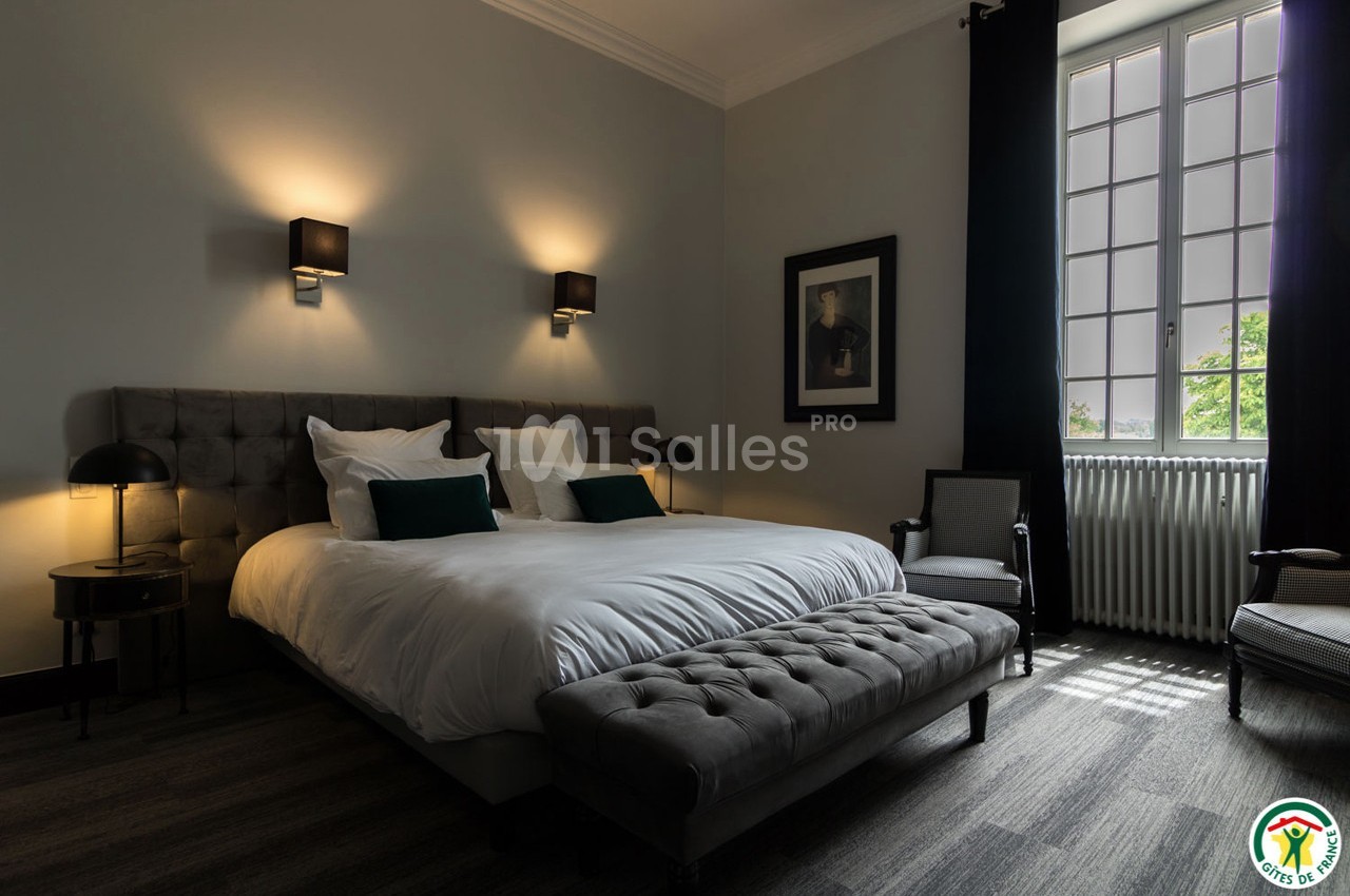 Chambre élégante avec lit double, éclairage tamisé, fauteuils et grande fenêtre laissant entrer la lumière naturelle.