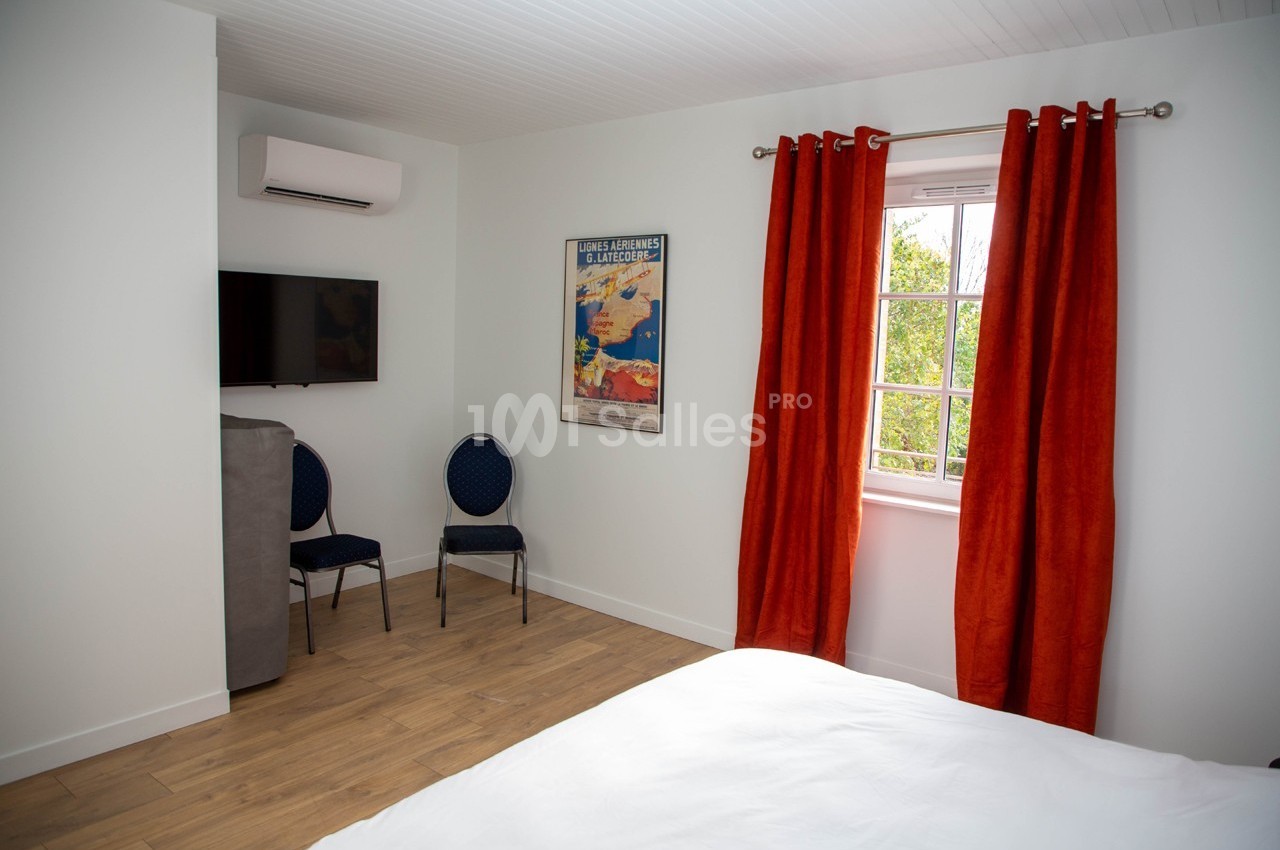 Chambre lumineuse avec un lit, un téléviseur mural, deux chaises, des rideaux rouges et une fenêtre donnant sur des arbres.