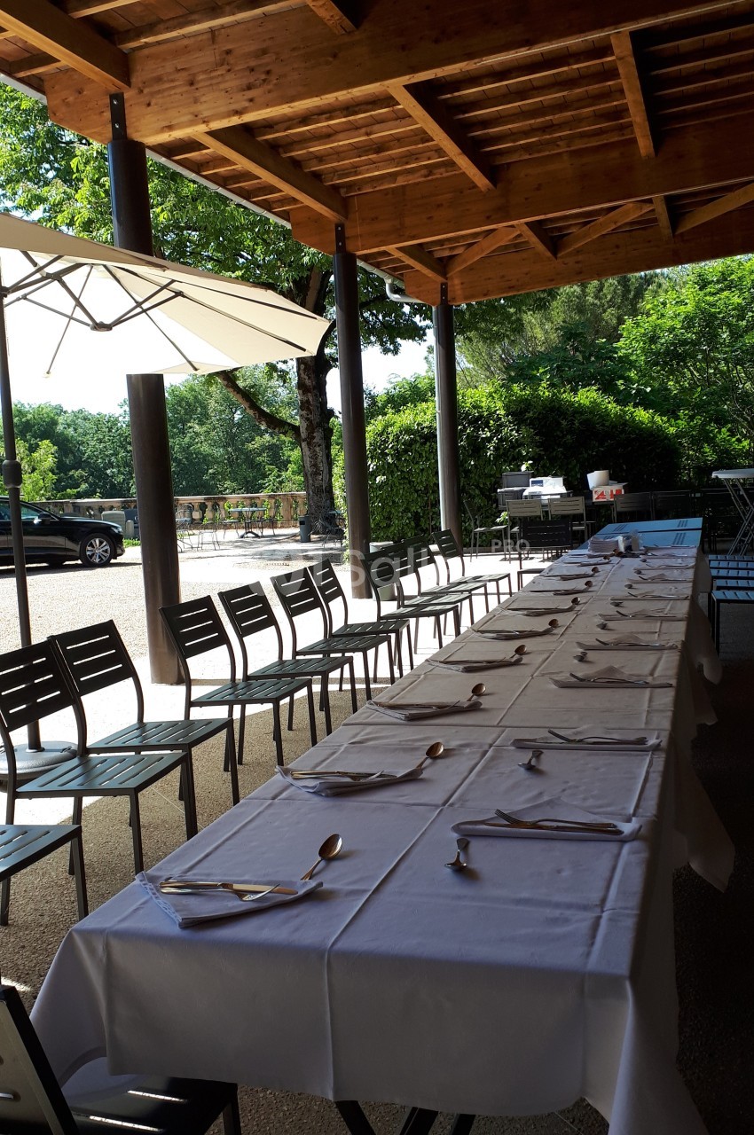 Tables alignées avec nappes blanches et couverts, disposées en extérieur sous une pergola en bois, entourées de chaises.