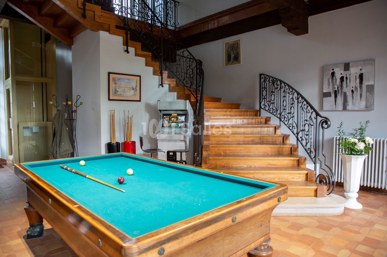 Table de billard dans une pièce lumineuse avec escalier en bois, décorations murales et porte-cannes à proximité.