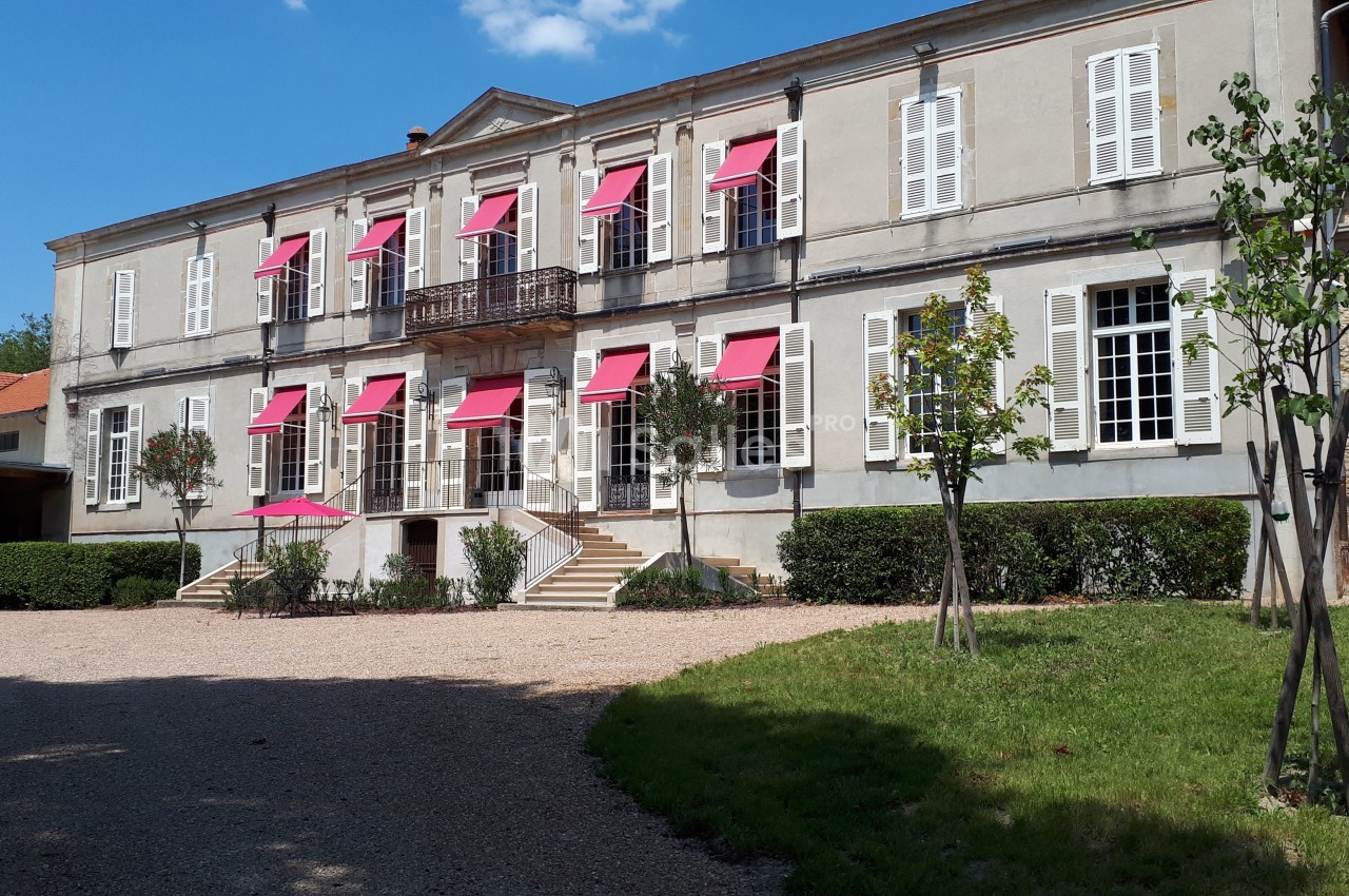 Façade d'un bâtiment ancien avec volets blancs et stores rouges, entouré d'arbres et d'une allée gravillonnée.