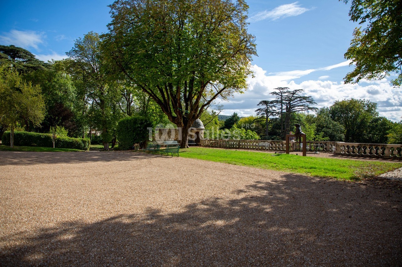 Allée gravillonnée bordée d'arbres et de verdure, avec bancs et balustrade en pierre sous un ciel partiellement nuageux.