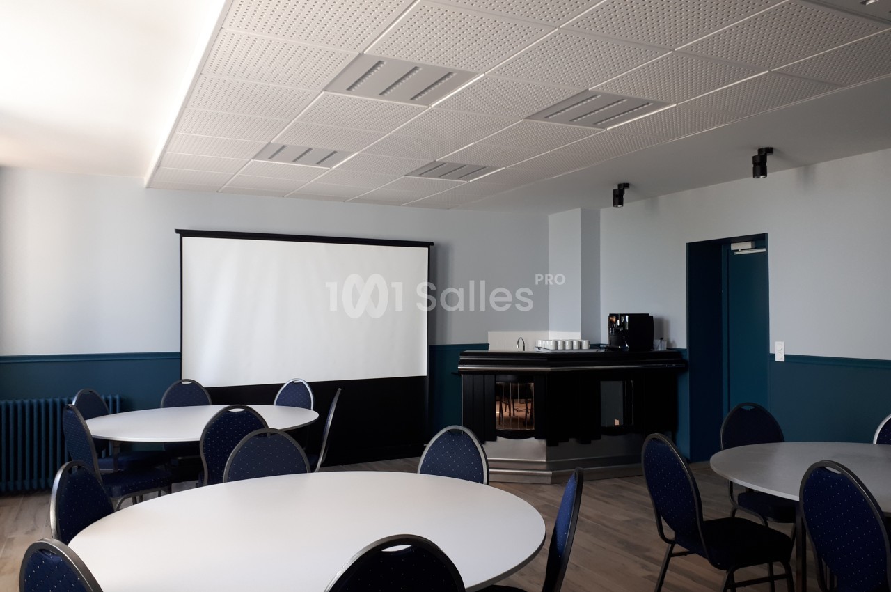 Salle de réunion avec tables rondes, chaises, écran de projection et coin café sur fond de murs clairs.