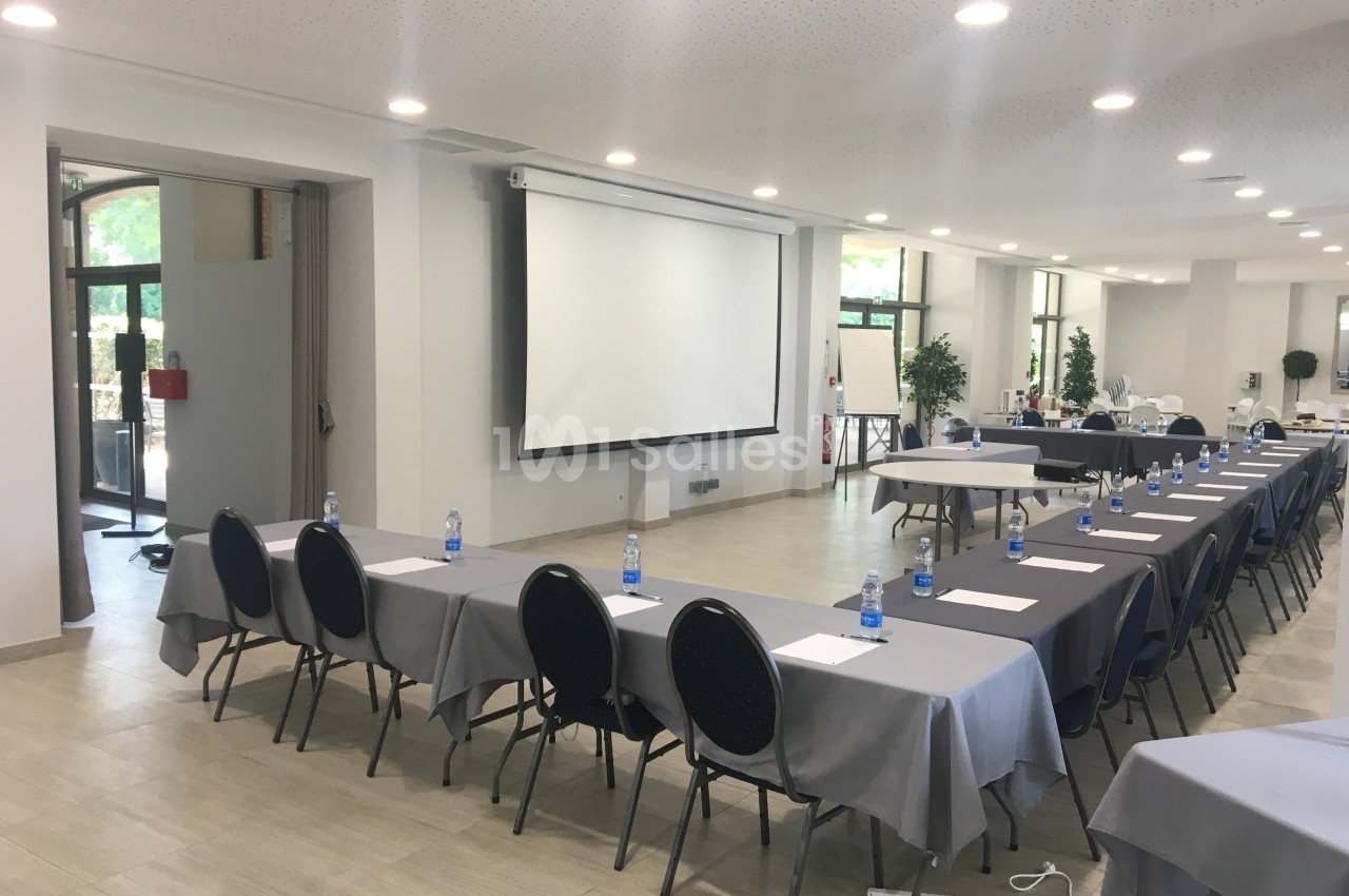 Salle de conférence lumineuse avec tables alignées, chaises, bouteilles d'eau et écran de projection.
