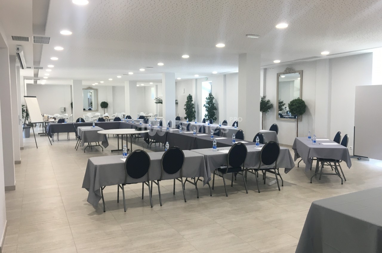 Salle de réunion lumineuse avec des tables rectangulaires couvertes de nappes grises, chaises noires et bouteilles d'eau.