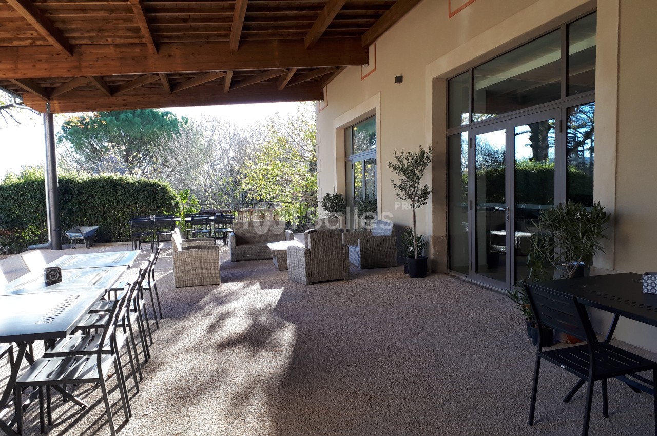 Terrasse couverte avec tables, chaises et fauteuils en rotin, entourée de verdure et baignée de lumière naturelle.