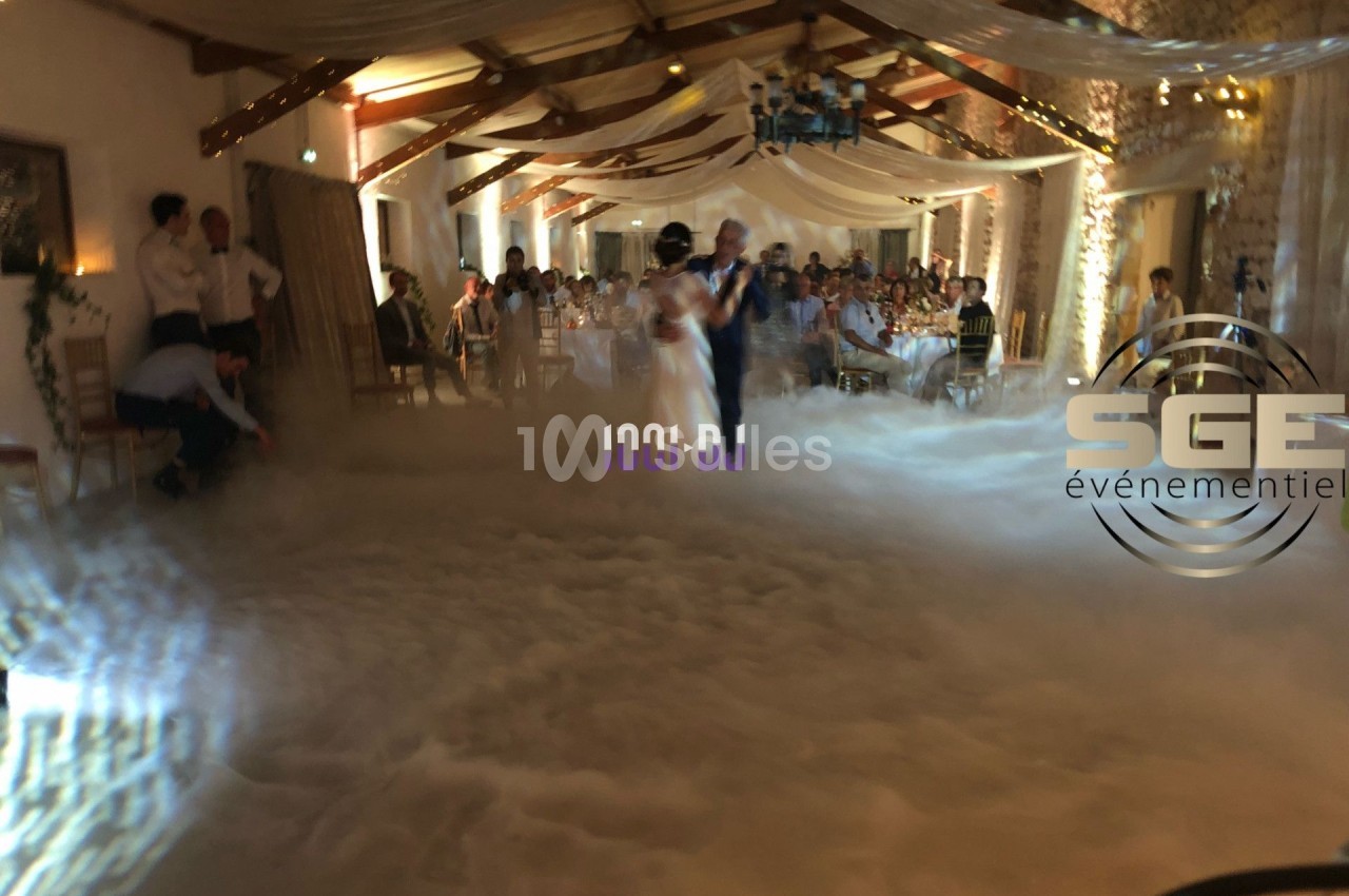 Un couple danse dans une salle de réception décorée, entourée de fumée au sol et d'invités assis.