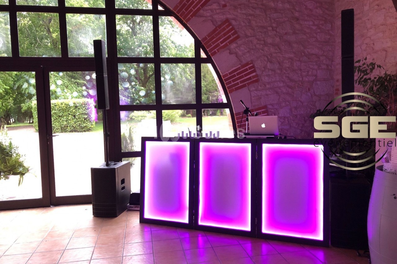 Poste de DJ avec éclairage LED violet, installé dans une salle avec murs en pierre et grande baie vitrée donnant sur un…
