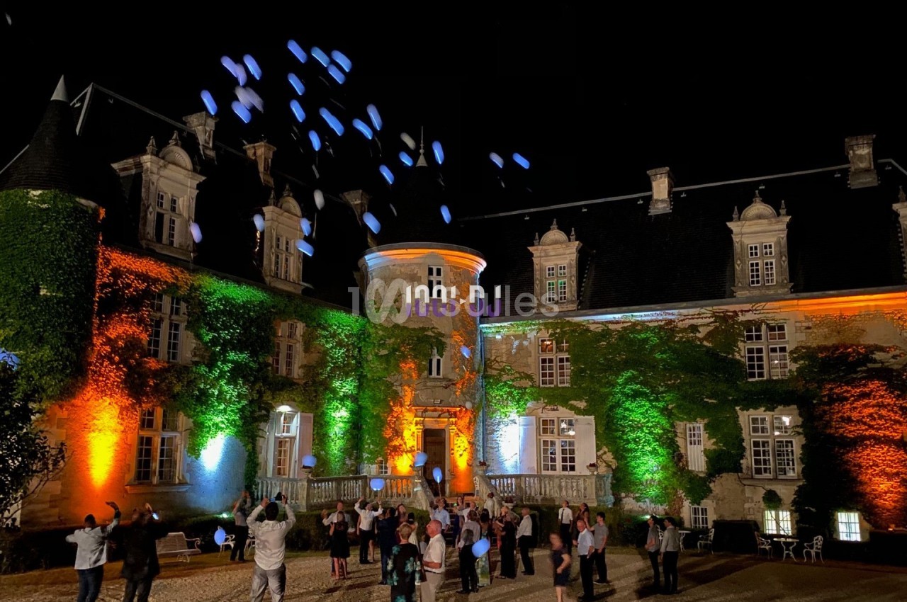 Des personnes libèrent des ballons lumineux devant un château éclairé de nuit par des projecteurs colorés.