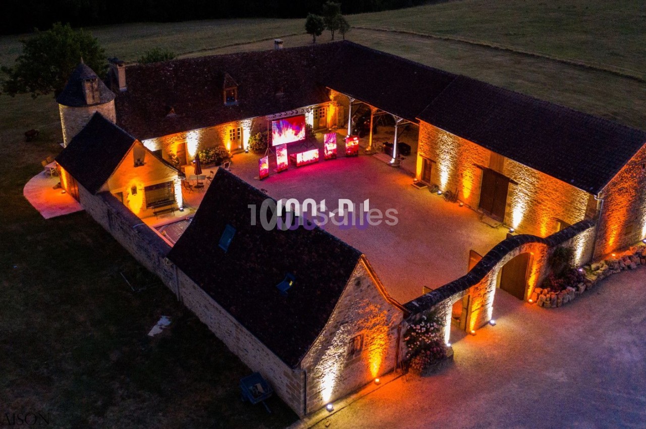 Vue aérienne d'une grande bâtisse en pierre éclairée par des lumières chaudes, entourée de pelouses.