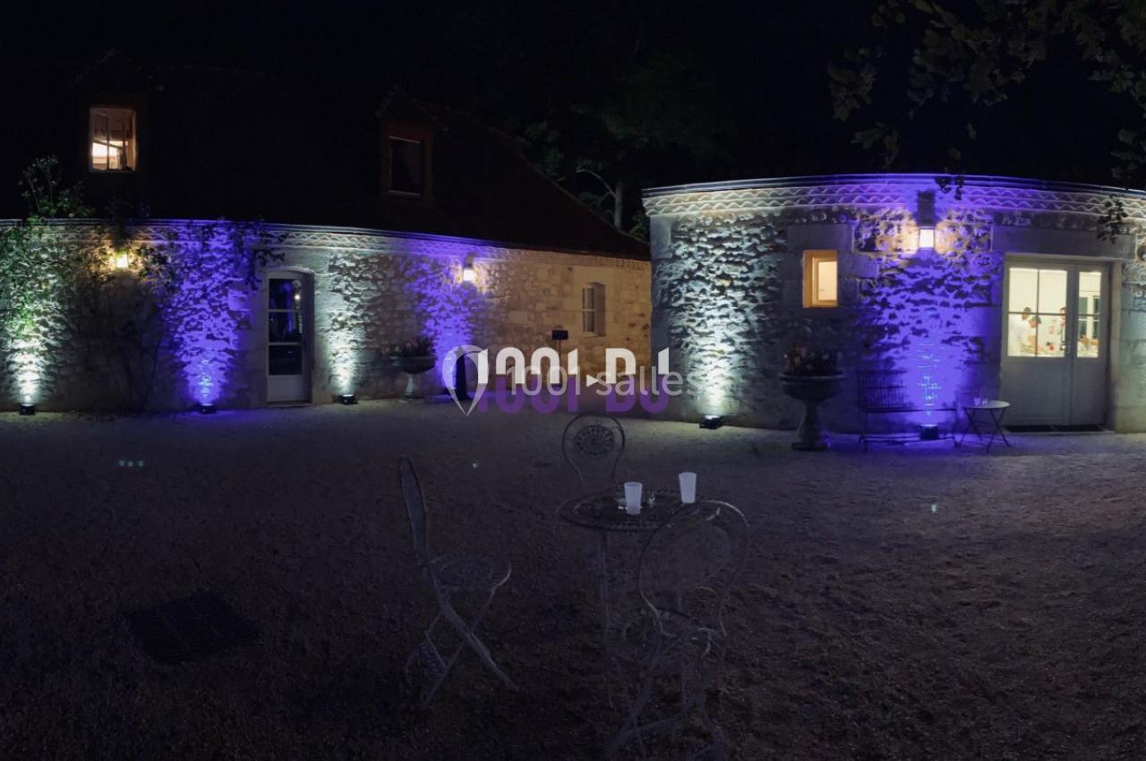 Façade d'un bâtiment en pierre éclairée par des lumières colorées, avec des tables et chaises en métal au premier plan.