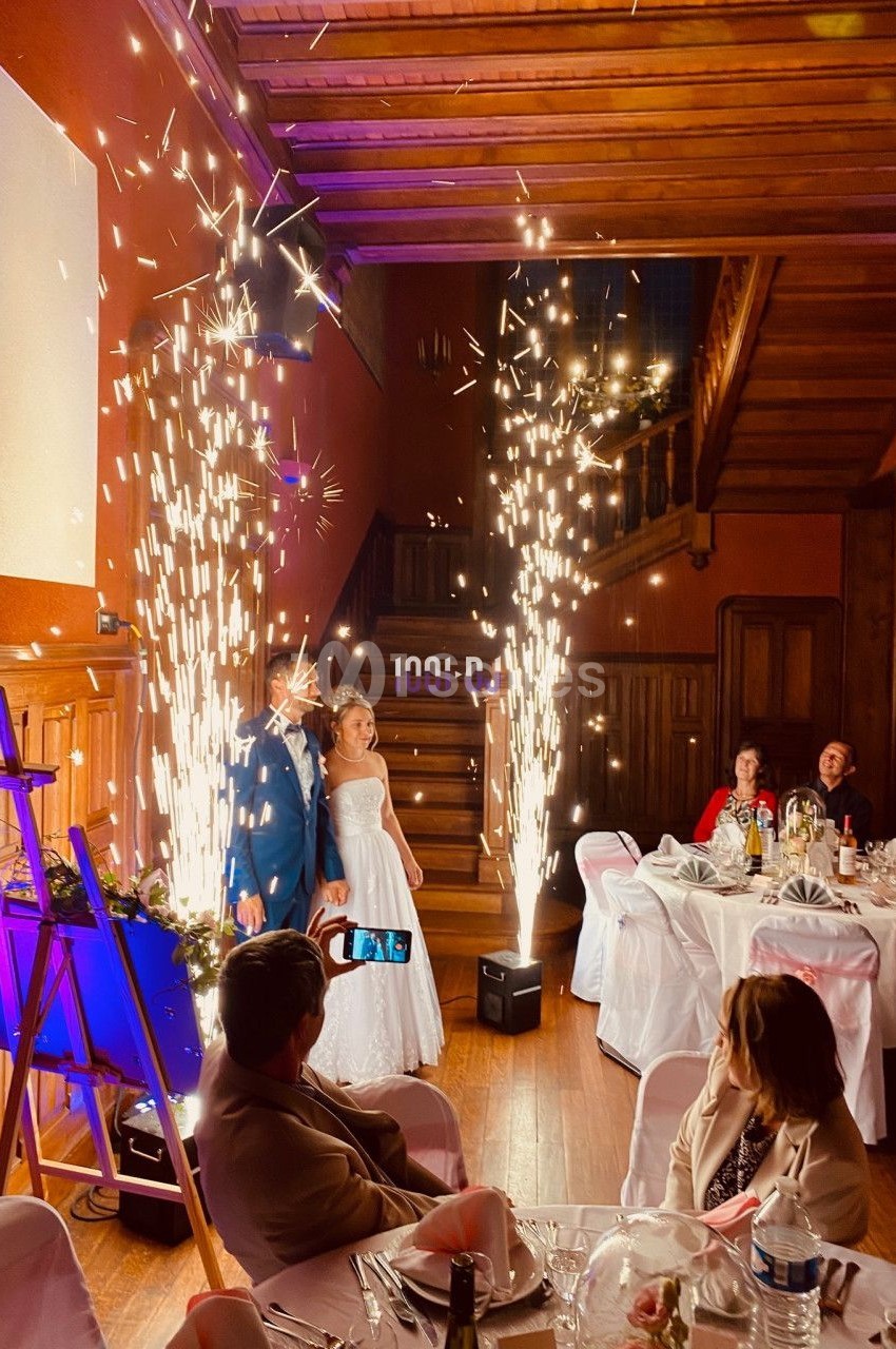 Un couple en tenue de mariage pose devant des fontaines d'étincelles dans une salle décorée avec des invités assis.