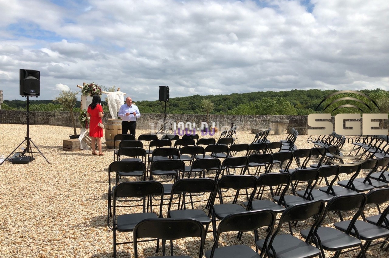 Chaises noires disposées en rangées sur un sol en gravier, avec une arche fleurie et des haut-parleurs en arrière-plan.