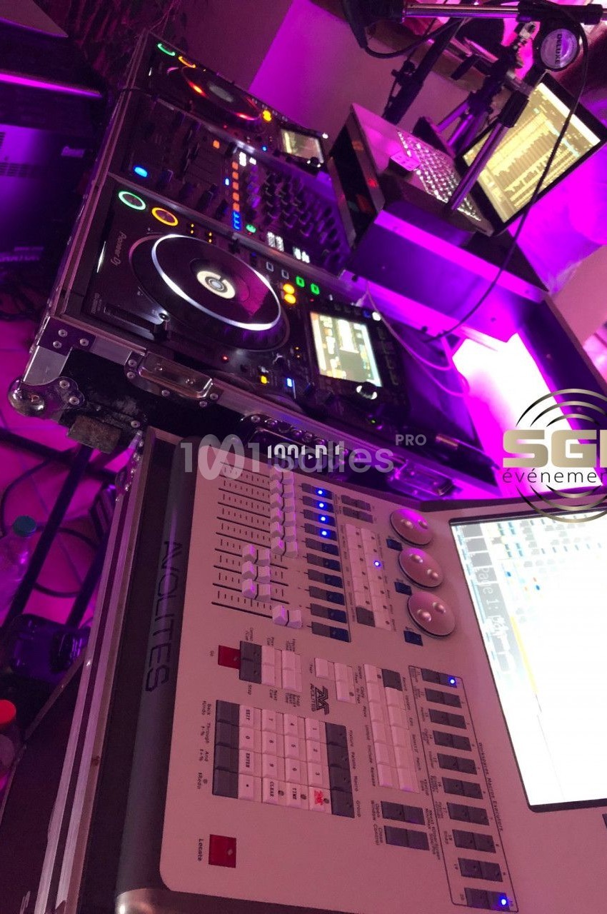 Matériel de DJ et console d'éclairage sur une table, entourés d'équipements techniques dans une ambiance lumineuse violette.