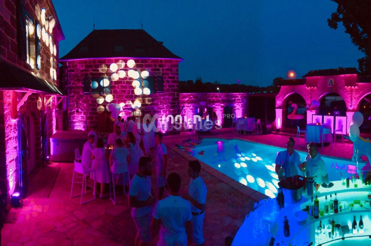 Soirée en extérieur près d'une piscine éclairée par des lumières colorées, avec des invités en tenue blanche.