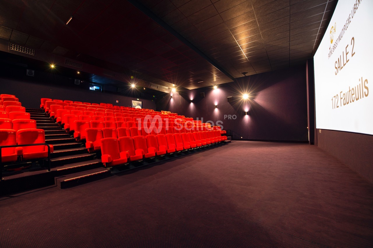Salle de cinéma vide avec rangées de fauteuils rouges, écran blanc et éclairage tamisé.
