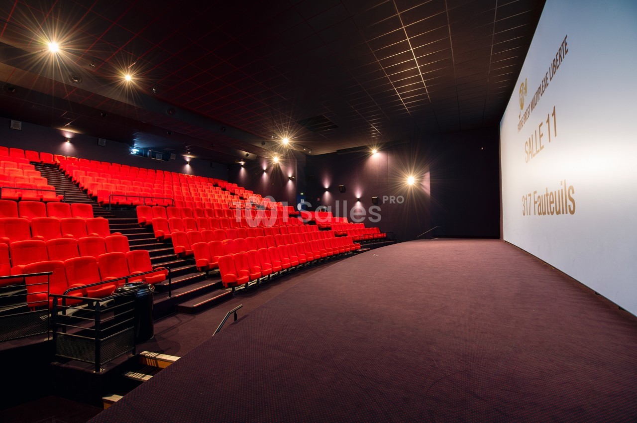 Salle de cinéma vide avec rangées de fauteuils rouges, écran blanc et éclairage tamisé.