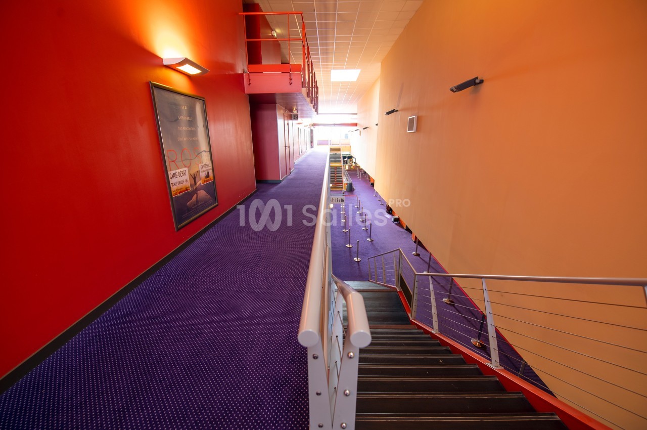 Couloir d'un cinéma avec moquette violette, murs rouges et jaunes, escalier en métal et affiches murales.