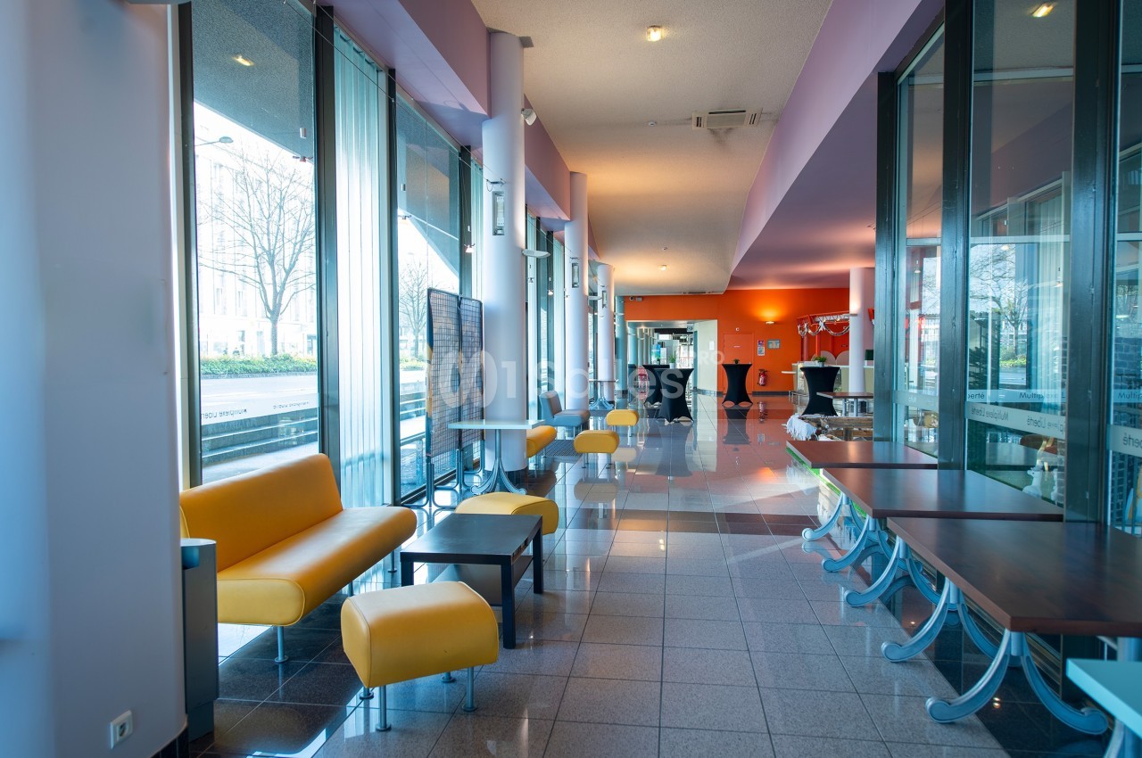 Espace intérieur lumineux avec canapés jaunes, tables et grandes baies vitrées donnant sur l'extérieur.