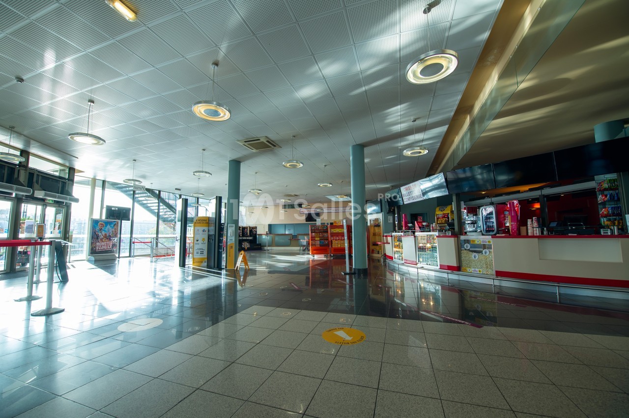 Hall d'entrée d'un cinéma avec comptoirs de vente, sol carrelé et lumière naturelle entrant par de grandes fenêtres.