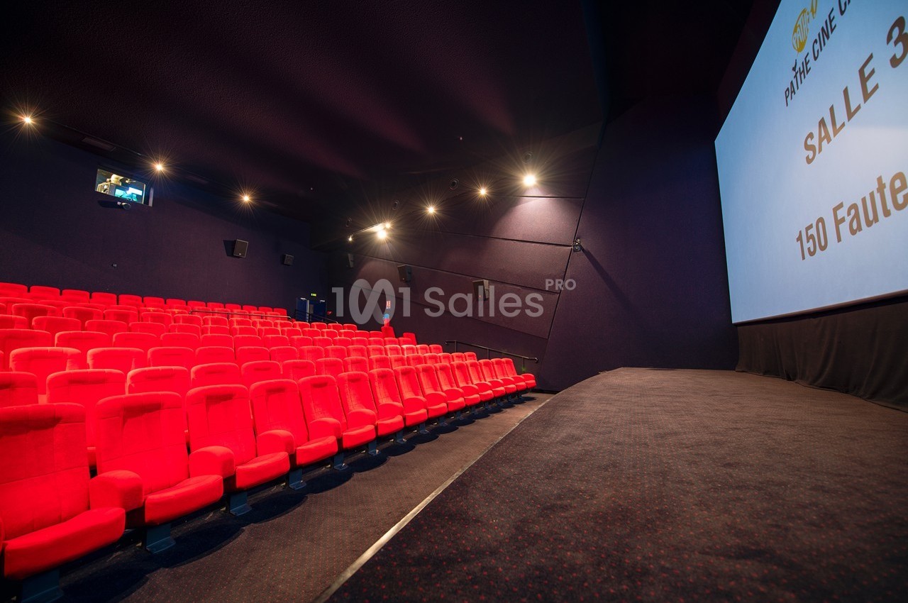Salle de cinéma vide avec rangées de fauteuils rouges, écran blanc à droite et éclairage tamisé.