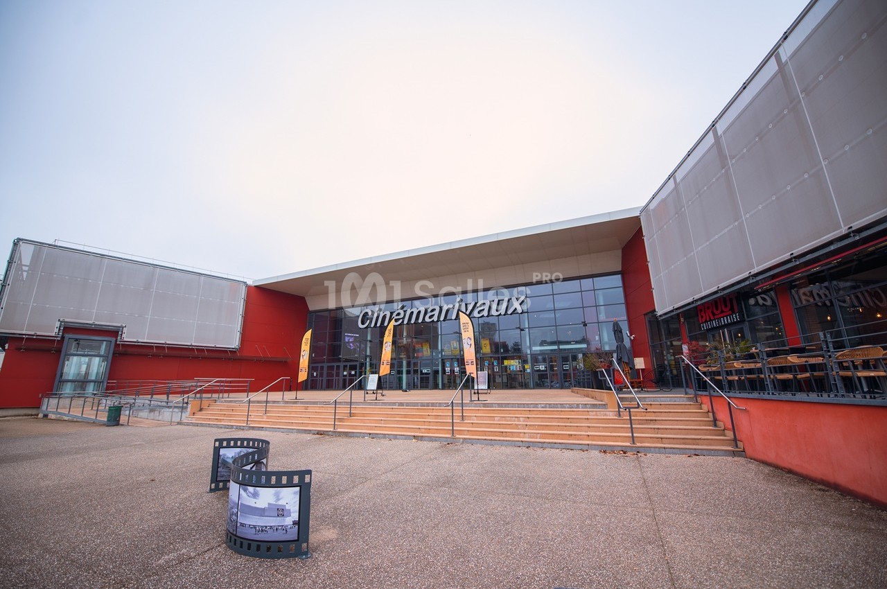 Façade d'un cinéma moderne avec entrée vitrée, escaliers et bannière publicitaire sur le parvis.