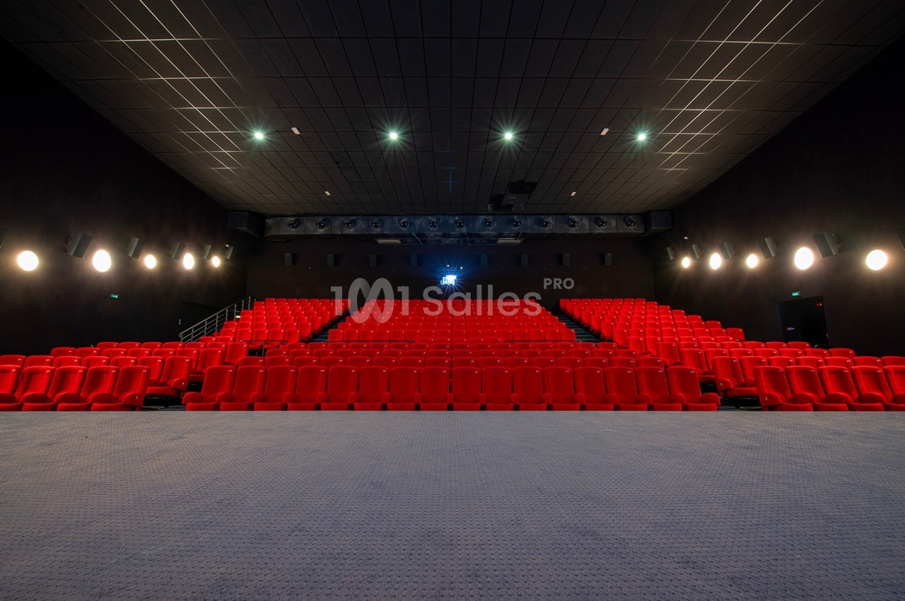Salle de cinéma vide avec des rangées de sièges rouges, éclairage tamisé et écran éteint au fond.