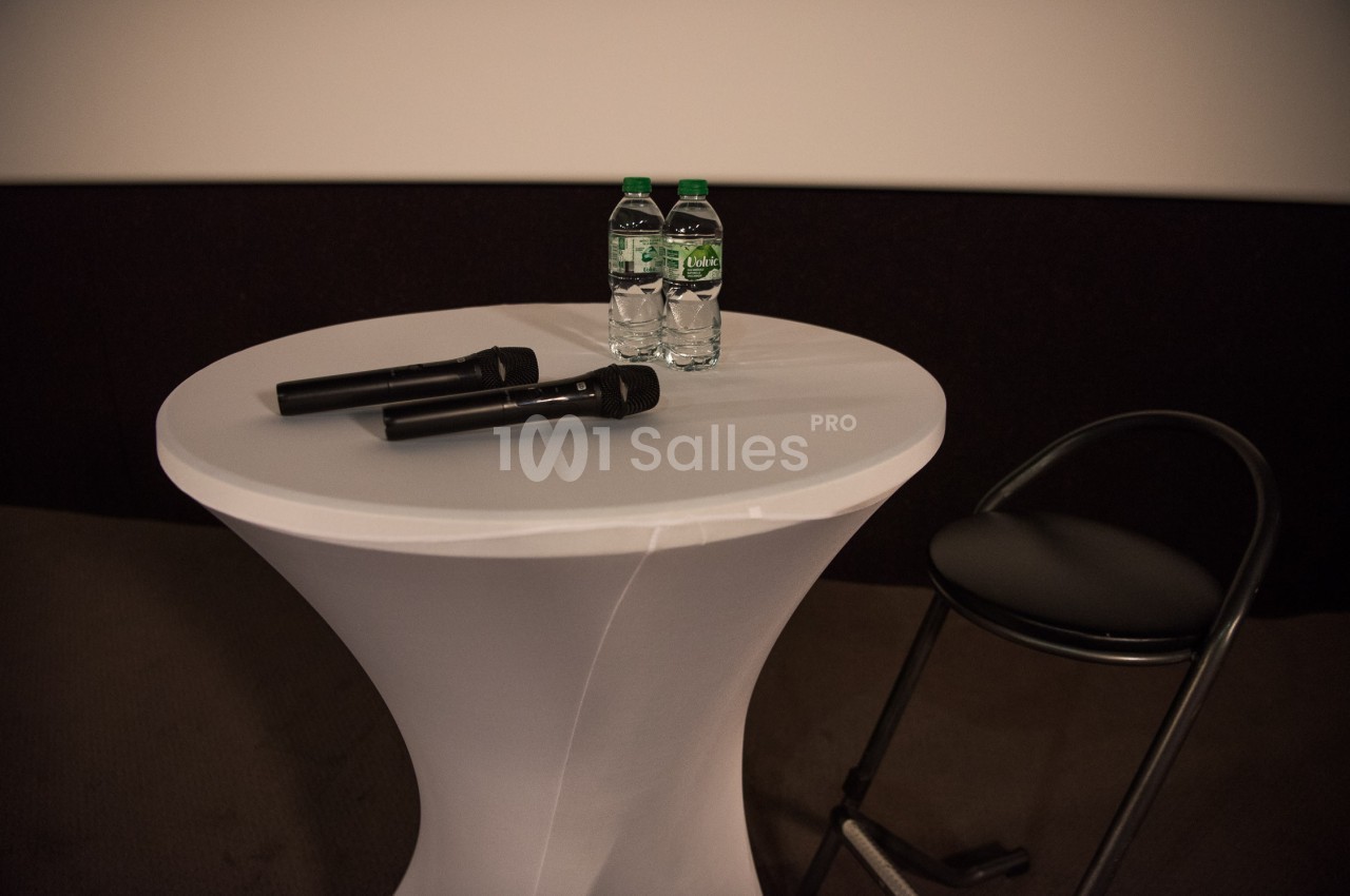 Deux microphones et deux bouteilles d'eau posés sur une table blanche, avec une chaise noire à côté.