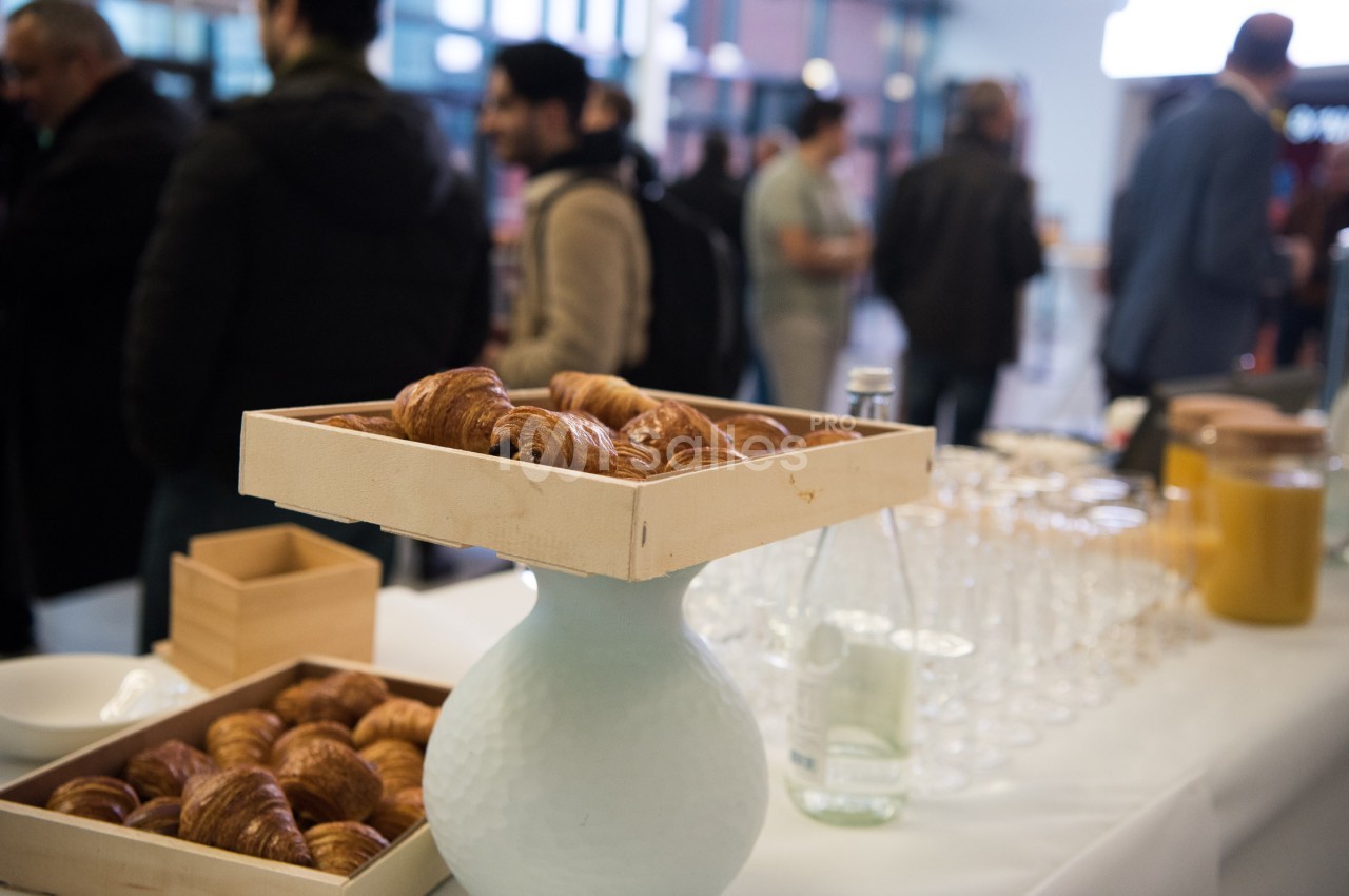 Plateau de croissants sur une table dressée avec des boissons et des verres, dans un espace avec des personnes en arrière…