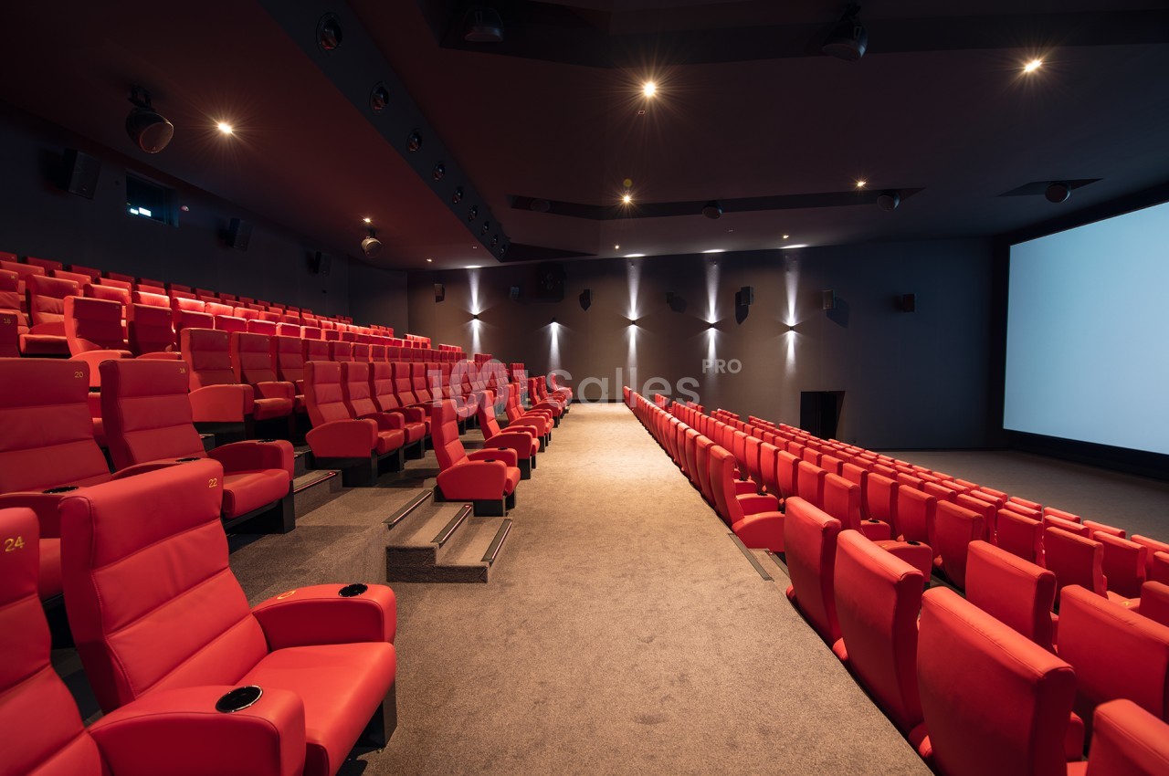 Salle de cinéma vide avec des rangées de fauteuils rouges et un grand écran blanc à droite.