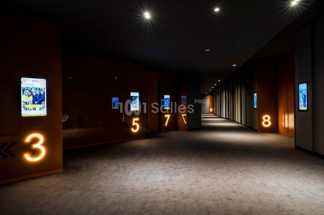Couloir sombre avec moquette, numéros lumineux sur les murs et affiches rétroéclairées alignées.