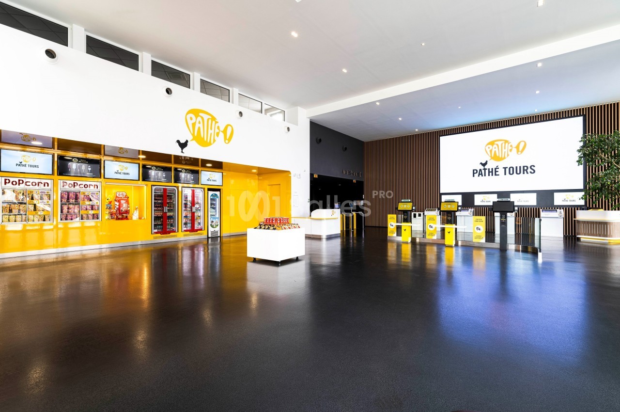 Hall d'entrée lumineux d'un cinéma avec comptoirs, distributeurs de snacks et écran affichant ’Pathé Tours’.