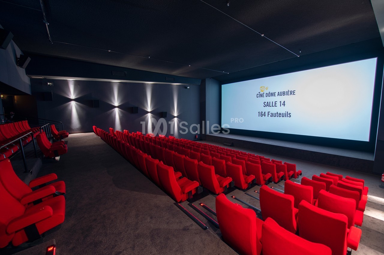 Salle de cinéma avec écran allumé, rangées de fauteuils rouges et éclairage tamisé.