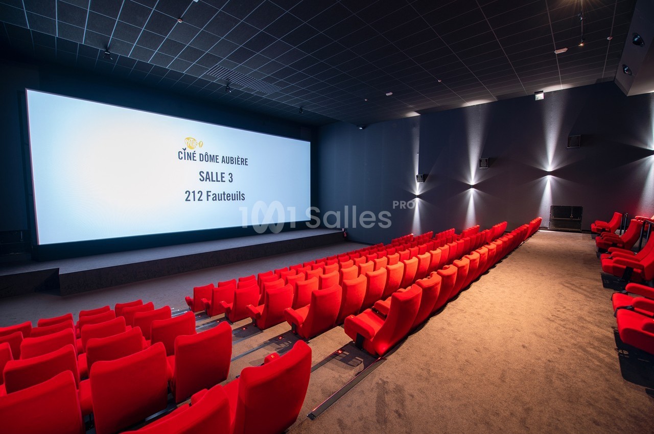 Salle de cinéma moderne avec rangées de fauteuils rouges, écran géant allumé et éclairage tamisé.