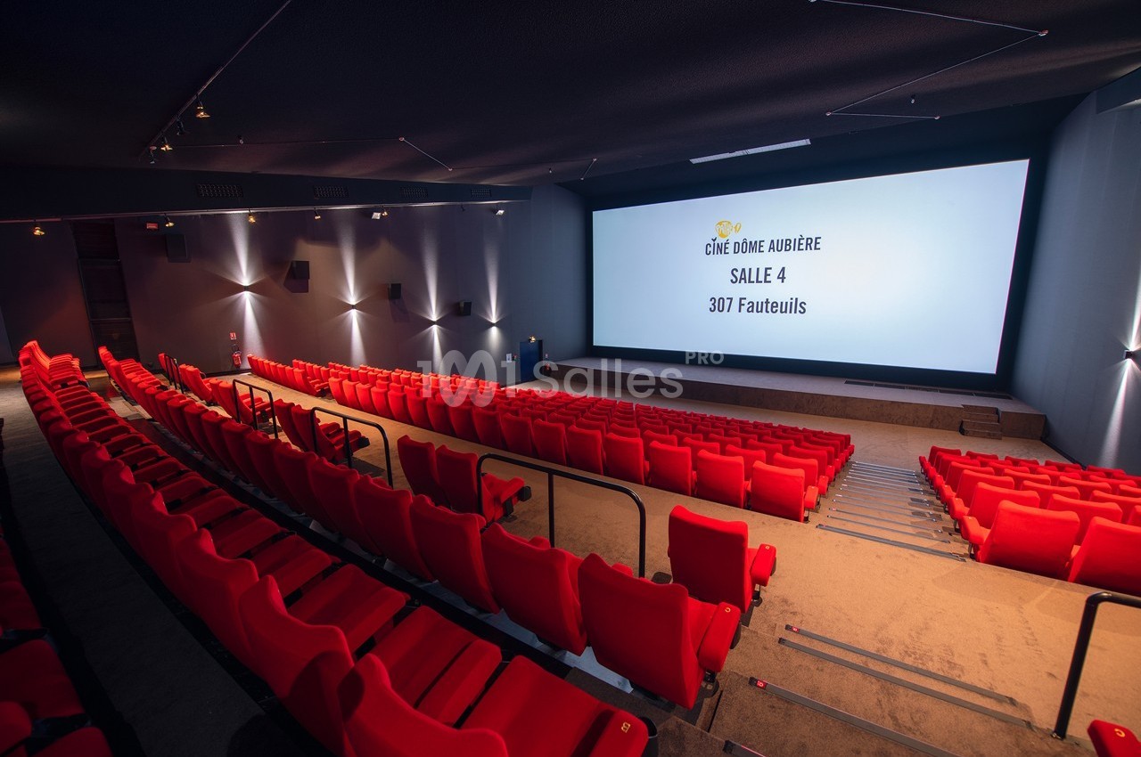 Salle de cinéma vide avec des sièges rouges, écran géant et éclairage tamisé.