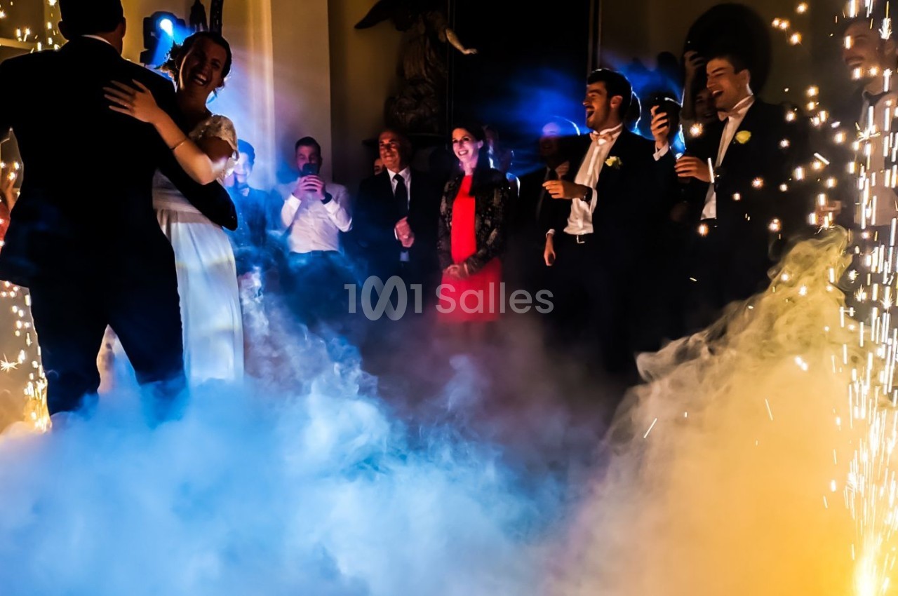 Un couple danse au milieu de la fumée et des étincelles, entouré d'invités élégants dans une ambiance festive.