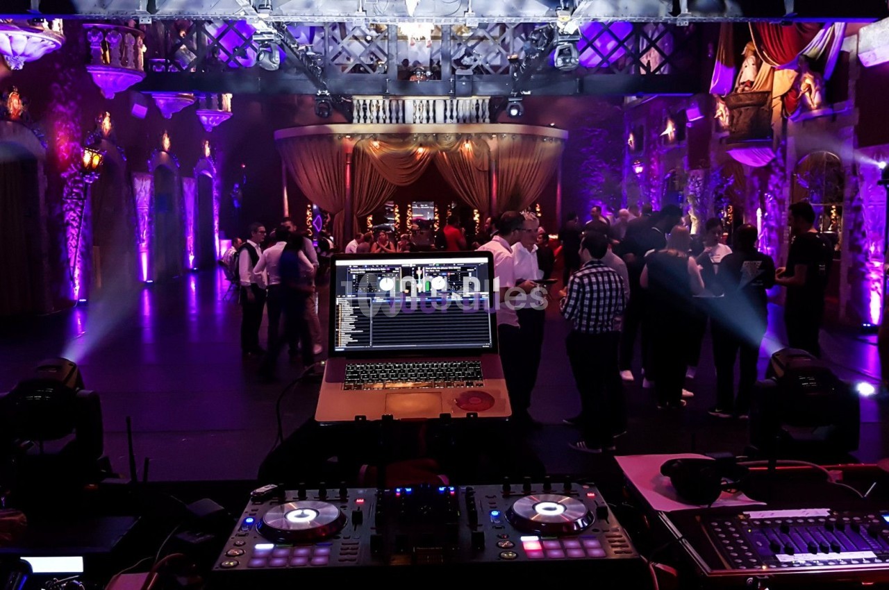 Vue depuis une table de mixage avec ordinateur, éclairages colorés et invités dansant dans une salle de réception.