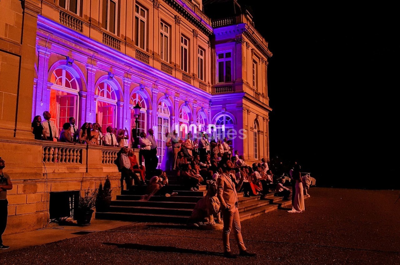 Façade d'un bâtiment illuminé en violet avec des personnes assises et debout sur les marches, de nuit.