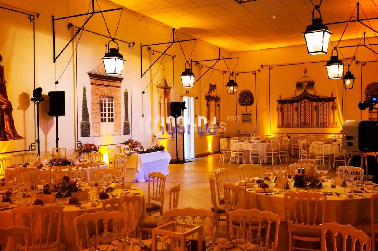 Salle de réception décorée avec des tables rondes dressées, des chaises en bois clair et des éclairages suspendus.