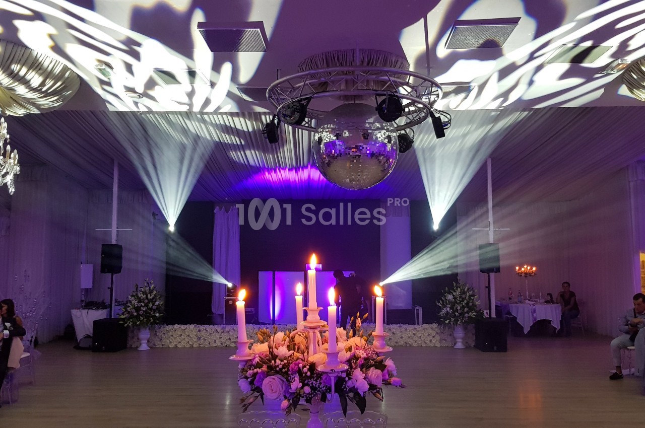 Salle de réception décorée avec des chandeliers, des fleurs et une boule disco, éclairée par des projecteurs.