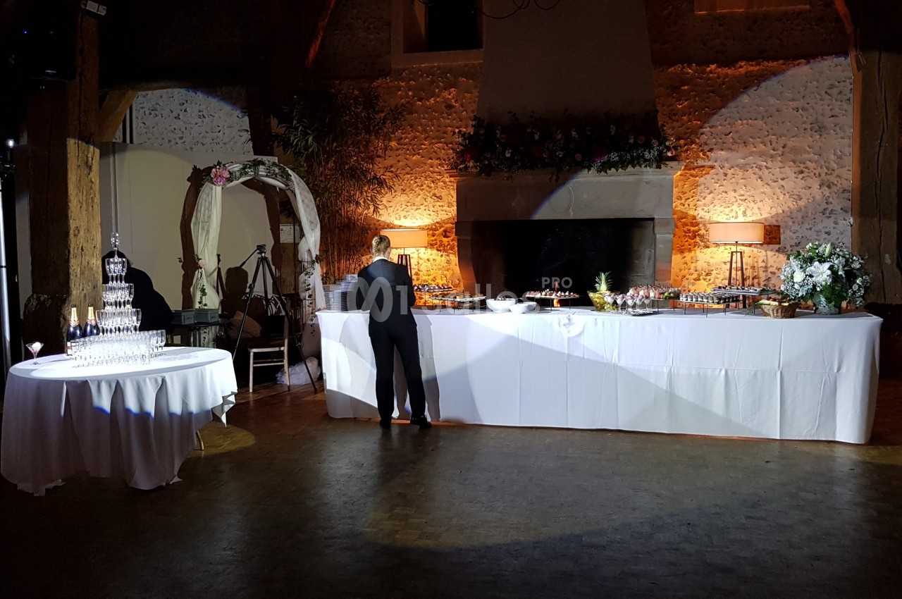 Salle de réception décorée avec une table de buffet éclairée, un présentoir à verres et une arche fleurie.