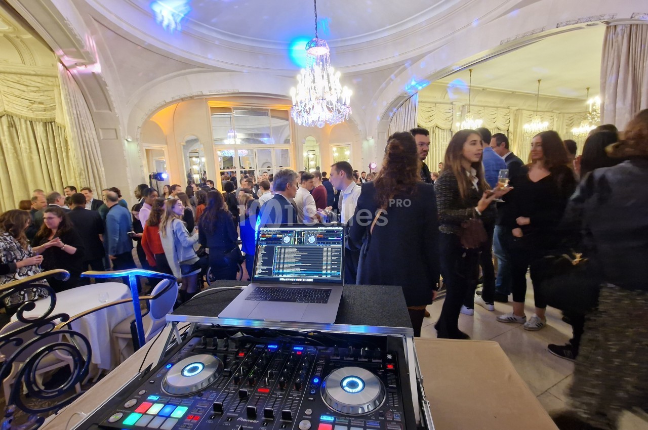 Vue d'une soirée dansante avec une table de mixage en premier plan et des invités dans une salle élégante en arrière-plan.