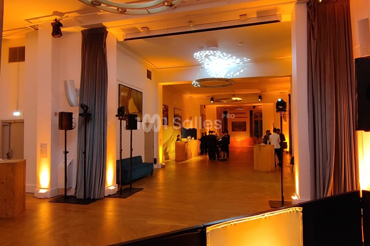 Salle de réception éclairée en orange avec des colonnes, des rideaux et des invités au fond près d'un comptoir.