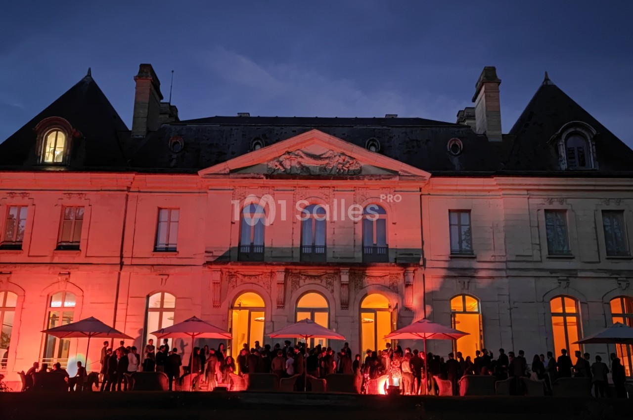 Façade d'un château éclairée en rouge à la tombée de la nuit, avec des invités rassemblés sous des parasols.