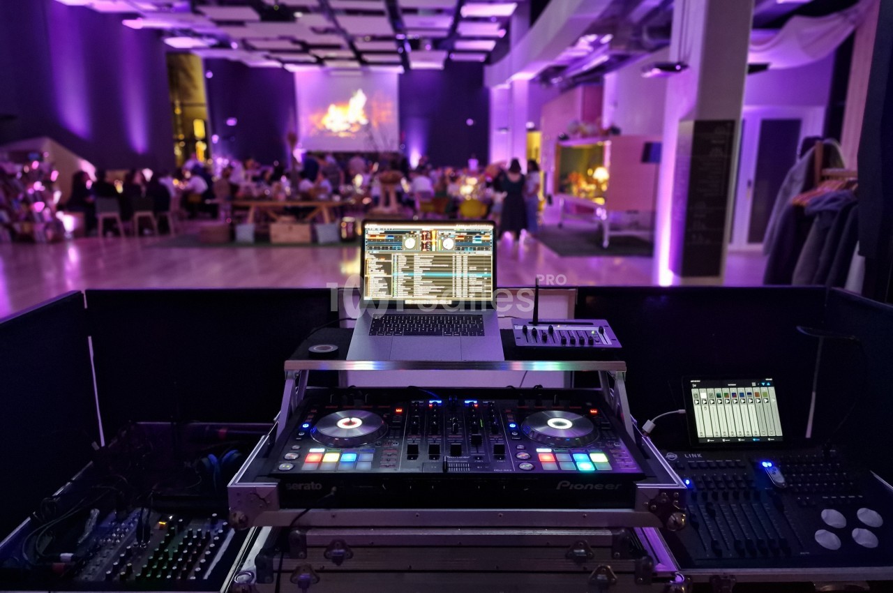 Vue d'une régie DJ avec platines et ordinateur, face à une salle éclairée en violet lors d'un événement festif.