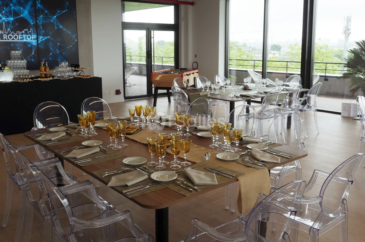 Salle lumineuse aménagée pour un événement, avec tables dressées, chaises transparentes et vue sur une terrasse extérieure.