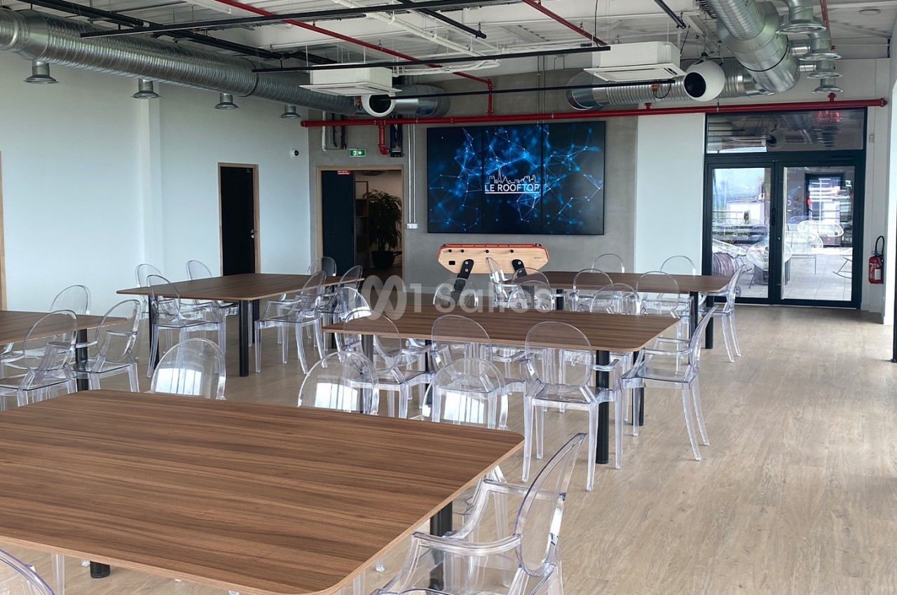 Salle lumineuse avec tables en bois, chaises transparentes, écran mural et baby-foot près de l'entrée vitrée.