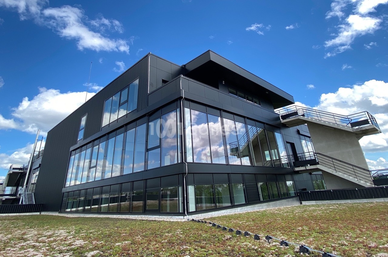 Bâtiment moderne à façade vitrée reflétant le ciel, entouré d'un toit végétalisé sous un ciel bleu avec nuages.