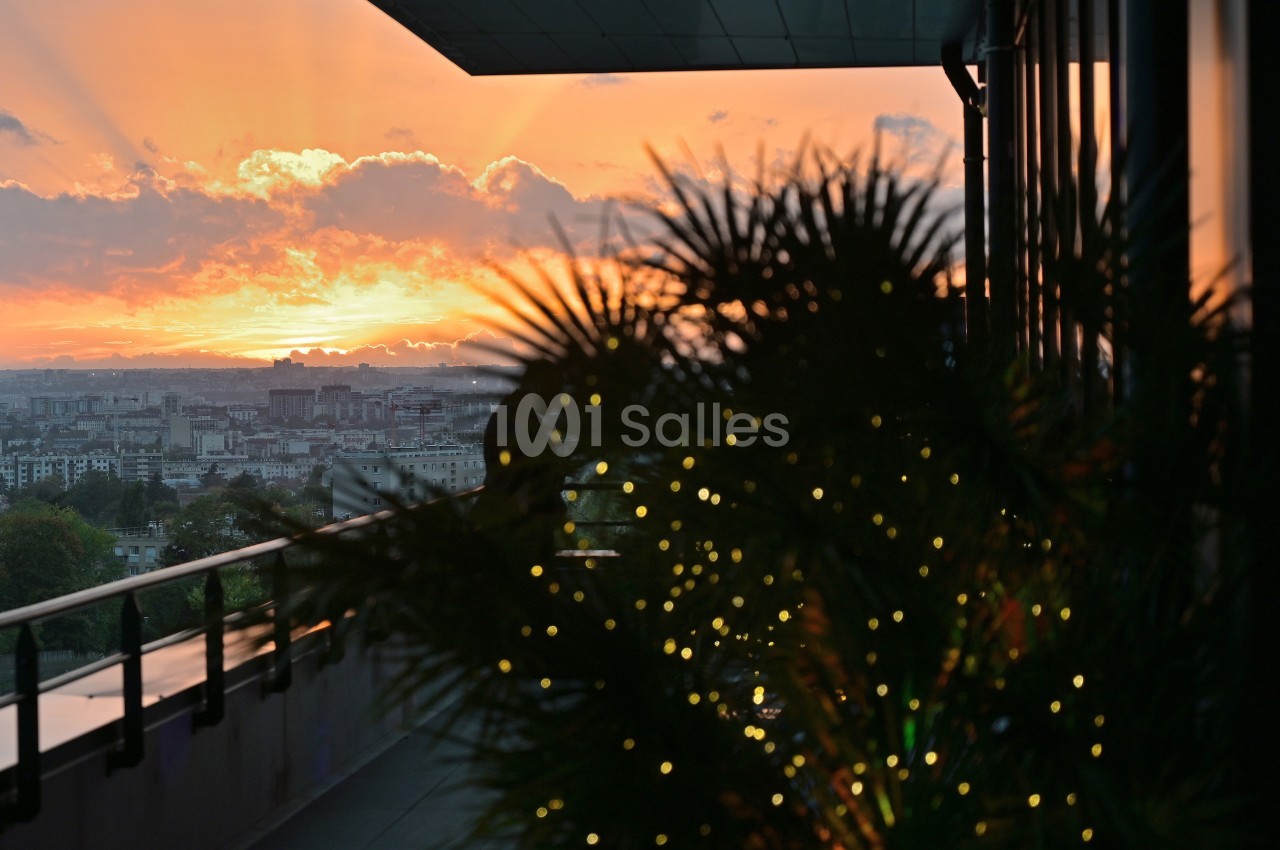 Coucher de soleil sur une ville, vu depuis un balcon avec des plantes illuminées au premier plan.
