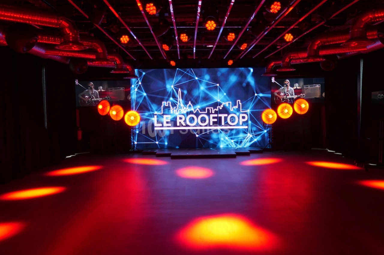 Salle sombre avec éclairage rouge, grand écran affichant ’Le Rooftop’ et décorations lumineuses suspendues.